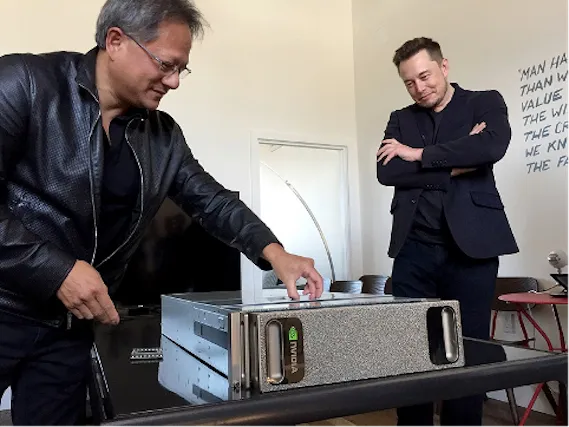 Jensen Huang in a black leather jacket sets a grey Nvidia server on a table while a man in a dark blazer looks on, arms crossed and smiling.