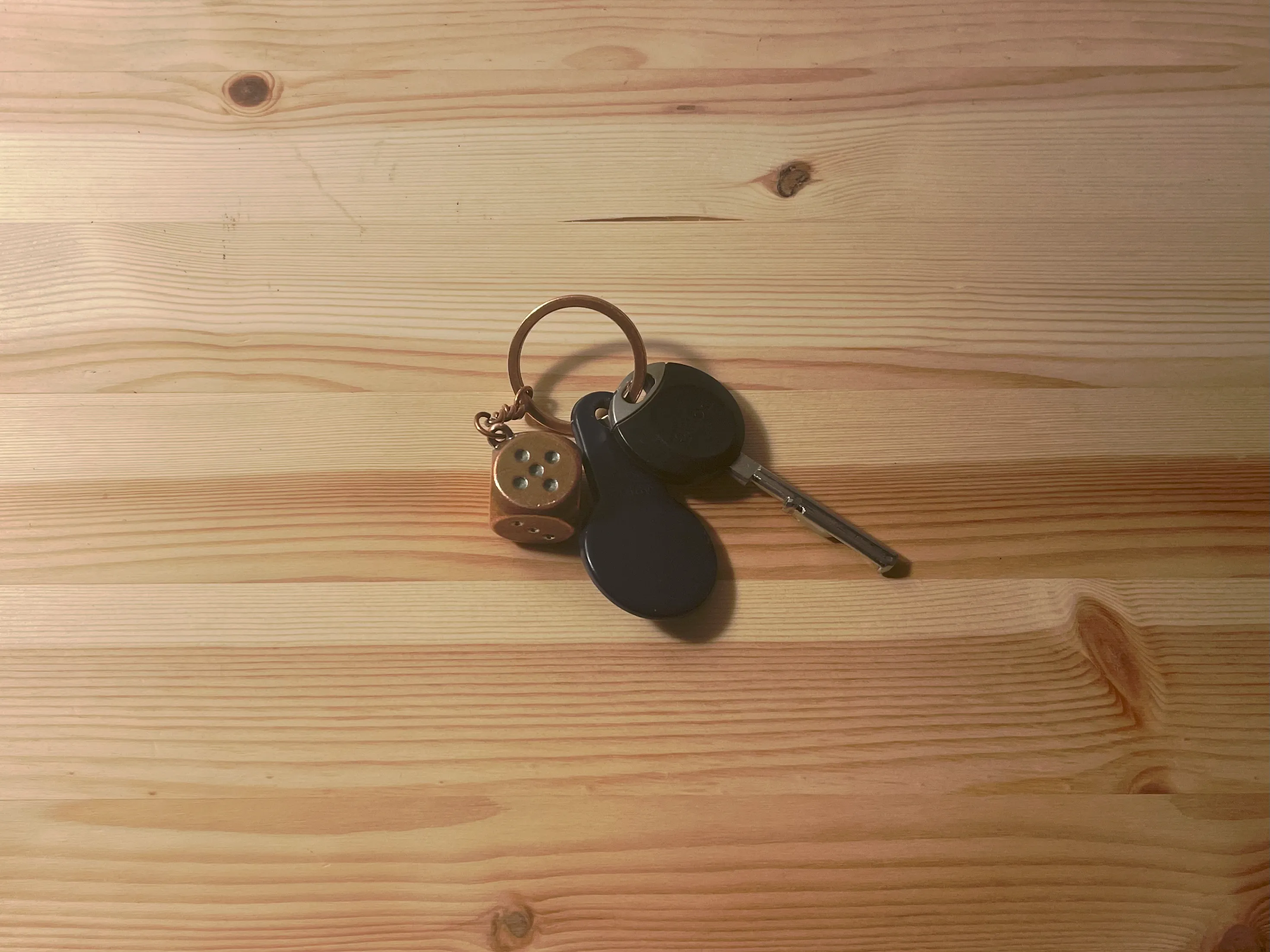 A keyring holding a small brown die, a dark oval fob and a single key, lying on a pale pine tabletop.
