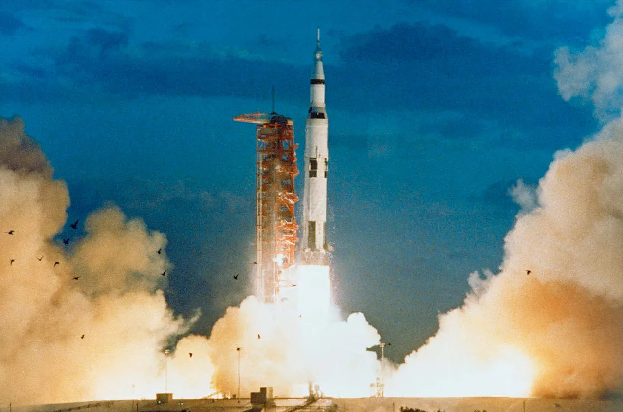 A Saturn V rocket rises from its launch pad beside a red tower, engines blazing amid billowing smoke under a dark blue sky.