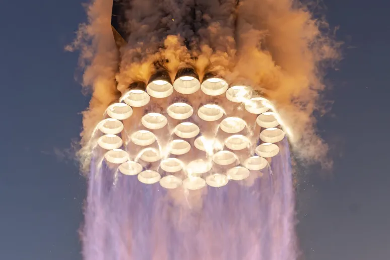View from below of Starship's Super Heavy booster in flight, its rings of Raptor engines glowing above violet exhaust and orange smoke.